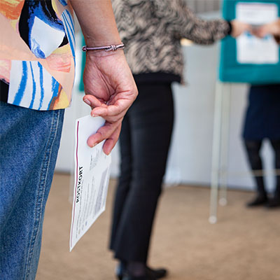 Närbild på en hand som håller i ett röstkort. I bakgrunden ses en vallokal.