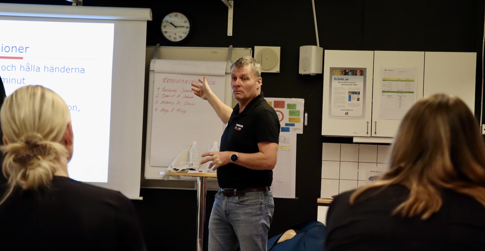 En man i 50-årsåldern står framför en whiteboard-tavla och instruerar elever i en lektionssal. 
