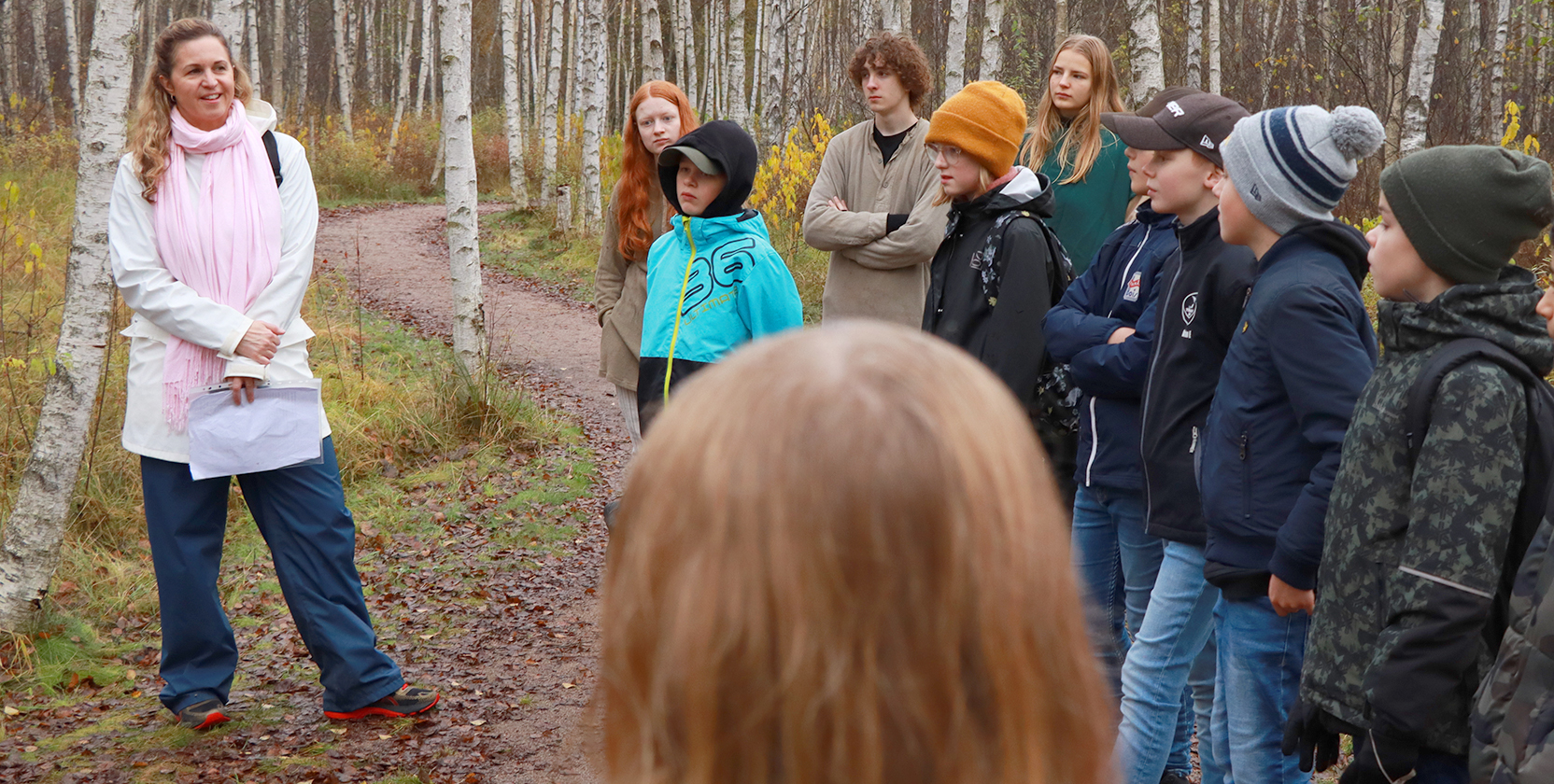 En lärare står med en grupp barn. Läraren står till vänster i bilden och tittar på eleverna. De är i en björkskog under hösten