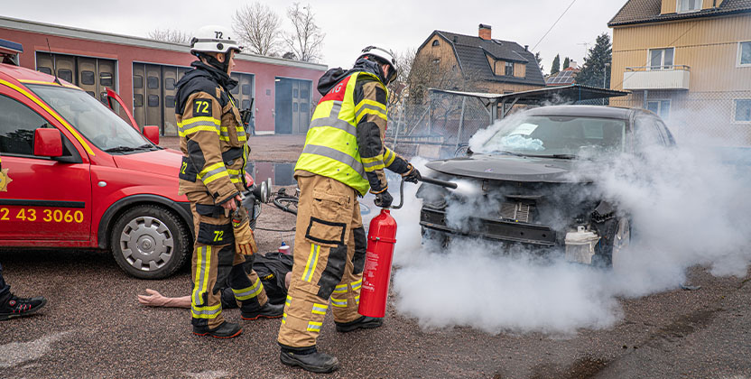 Två brandmän släcker en rykande bilbrand vid en olycksplats, med en röd brandbil i bakgrunden.