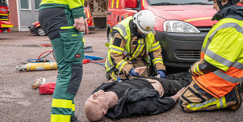 Räddningspersonal övar första hjälpen på en docka utomhus vid en röd utryckningsbil.