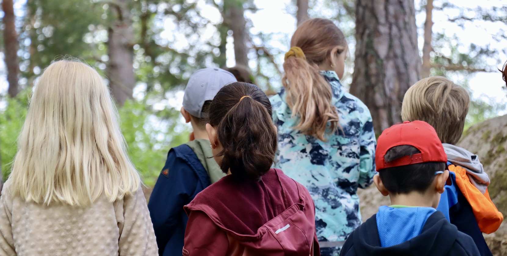 Barn i förskoleåldern, iklädda utomhuskläder, står tillsammans och lyssnar på två tjejer i 18-årsåldern utklädda till rumpnissar i skogen. 