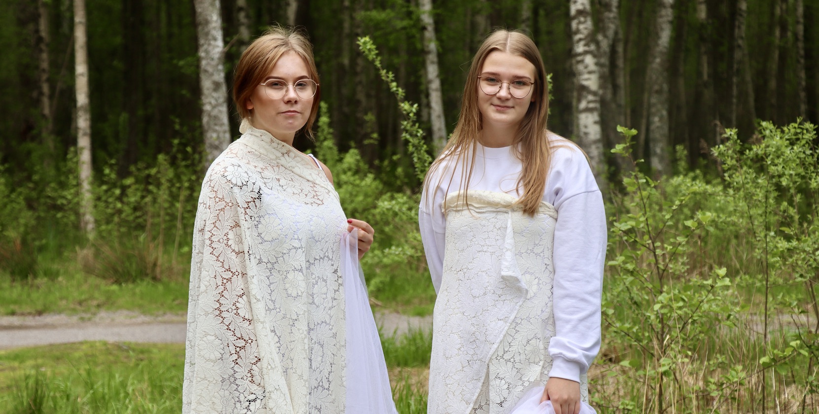 Två blonda tjejer i 18-årsåldern står på en öppen yta i skogen iklädda vita, fladdriga kläder - utklädda till älvor.