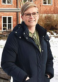 Camilla Amredahl har ljust kort hår, glasögon och står utanför en tegelbyggnad. Det är vinter och snö på marken.